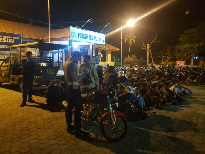 Teks: Puluhan pemuda dan kendaraan bermotor balap liar terjaring operasi.(blok-a.com/Syahrul Wijaya) Pewarta: Syahrul Wijaya Berita: Liputan Judul: Polsek Trowulan dan TNI Amankan 43 Motor dalam Razia Balap Liar Mojokerto, blok-a.com – Sebanyak 43 sepeda motor berhasil diamankan dalam razia balap liar yang digelar oleh Polsek Trowulan, TNI, dan Polres Mojokerto pada Minggu dini hari (22/9). Operasi gabungan ini bertujuan untuk menjaga keamanan dan ketertiban di wilayah hukum Polsek Trowulan. Operasi dimulai sekitar pukul 00.30 WIB dengan apel persiapan di depan Pendopo Kecamatan Trowulan. Kapolsek Trowulan, Kompol Margo Sukwandi, SH, memimpin apel tersebut dan memberikan arahan kepada anggotanya. Dalam instruksinya, petugas berpakaian preman diperintahkan masuk ke lokasi terlebih dahulu, sementara tim lainnya ditugaskan untuk menutup akses jalan di beberapa titik strategis, termasuk di jalan utama Dusun, serta simpul jalan di sekitar Jalan Raya Domas. Tim gabungan yang terdiri dari Polri, TNI, dan Satpol PP, dikerahkan ke beberapa wilayah seperti Dusun Sambigede, Brumbung, Temboro, Kasihan, dan Penewon. Penutupan jalan dilakukan di area selatan Dusun Penewon dan utara Desa Jambuwok, guna memblokir akses keluar para pelaku balap liar. "Sebanyak 50 orang pengendara terjaring dalam operasi ini, dan mereka diminta berjalan kaki menuju Polsek Trowulan," terangnya. Sementara itu, sepeda motor mereka diangkut menggunakan truk dan diamankan di Polres Mojokerto. "Semua kendaraan yang terlibat diberikan tindakan tilang sesuai dengan aturan yang berlaku," pungkasnya. Operasi ini melibatkan 39 personel gabungan yang terdiri dari; Kabag Ops Polres Mojokerto, Kompol Hendro Sosianto, S.H., Kapolsek Trowulan, Kompol Margo Sukwandi, S.H, 32 personel Polsek Trowulan dan Polres Mojokerto, 6 personel TNI, 1 personel Satpol PP. Selama pelaksanaan operasi, situasi dilaporkan berjalan aman dan kondusif tanpa adanya insiden yang mengganggu.(Sya)